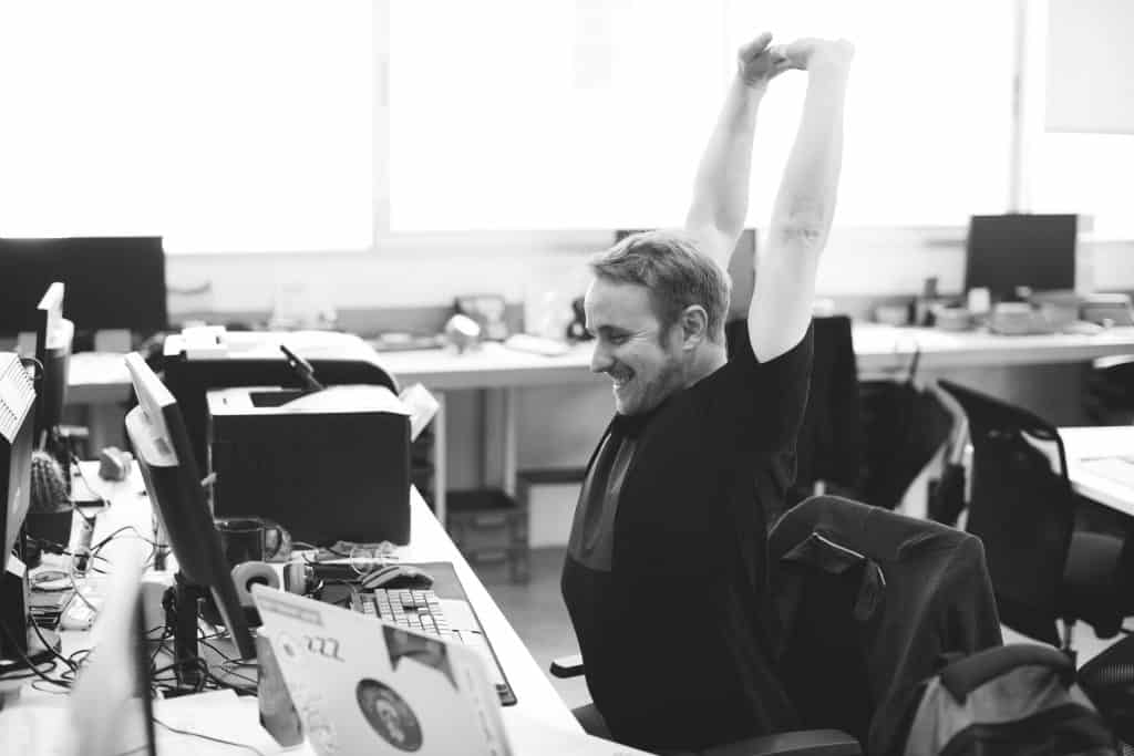 A man stretching and smiling at his desk, feeling accomplished in the open office setting. 